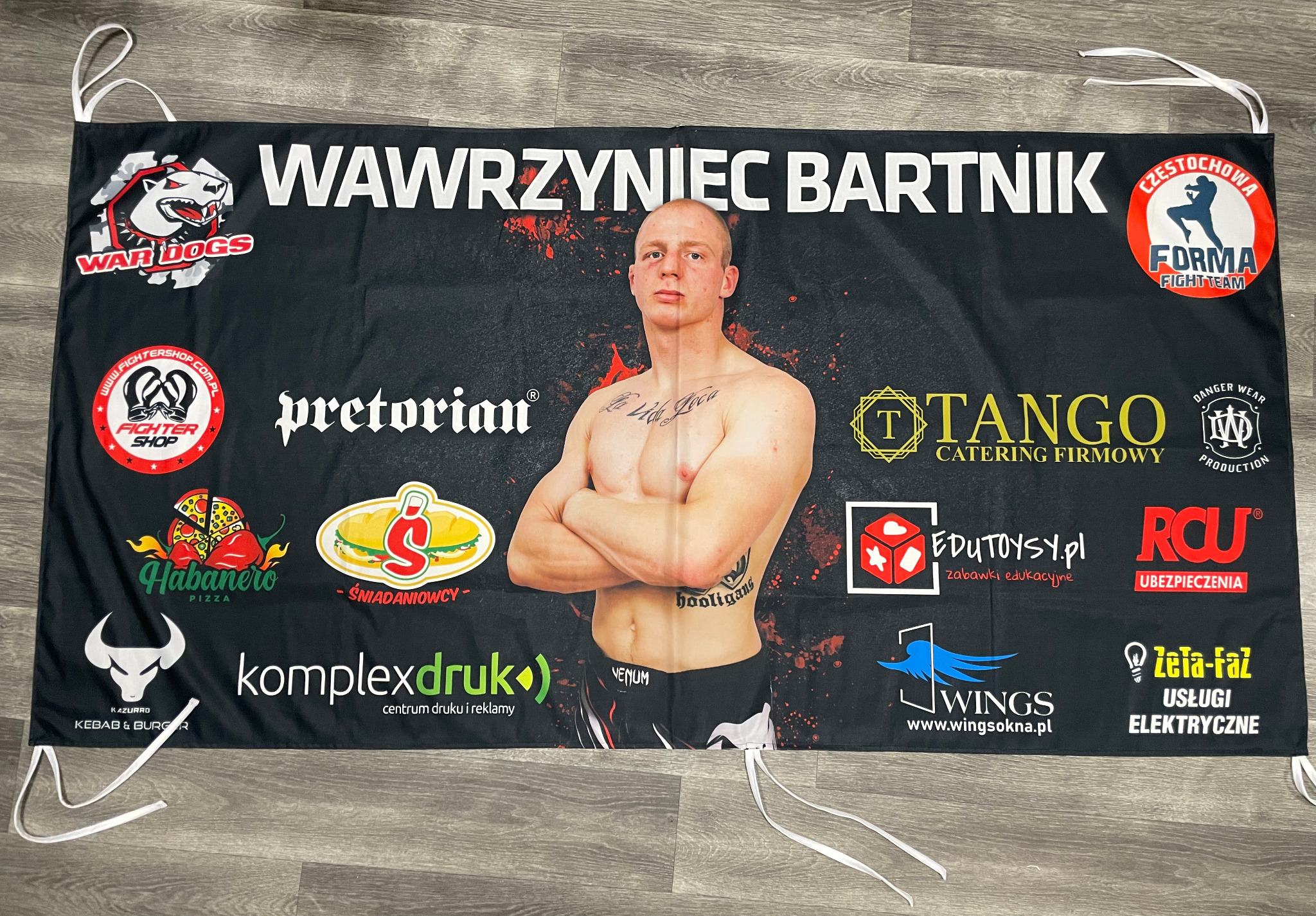 Baner reklamowy z wizerunkiem zawodnika sportów walki, logotypami sponsorów i nazwą klubu sportowego, zawieszony na sznurkach.
