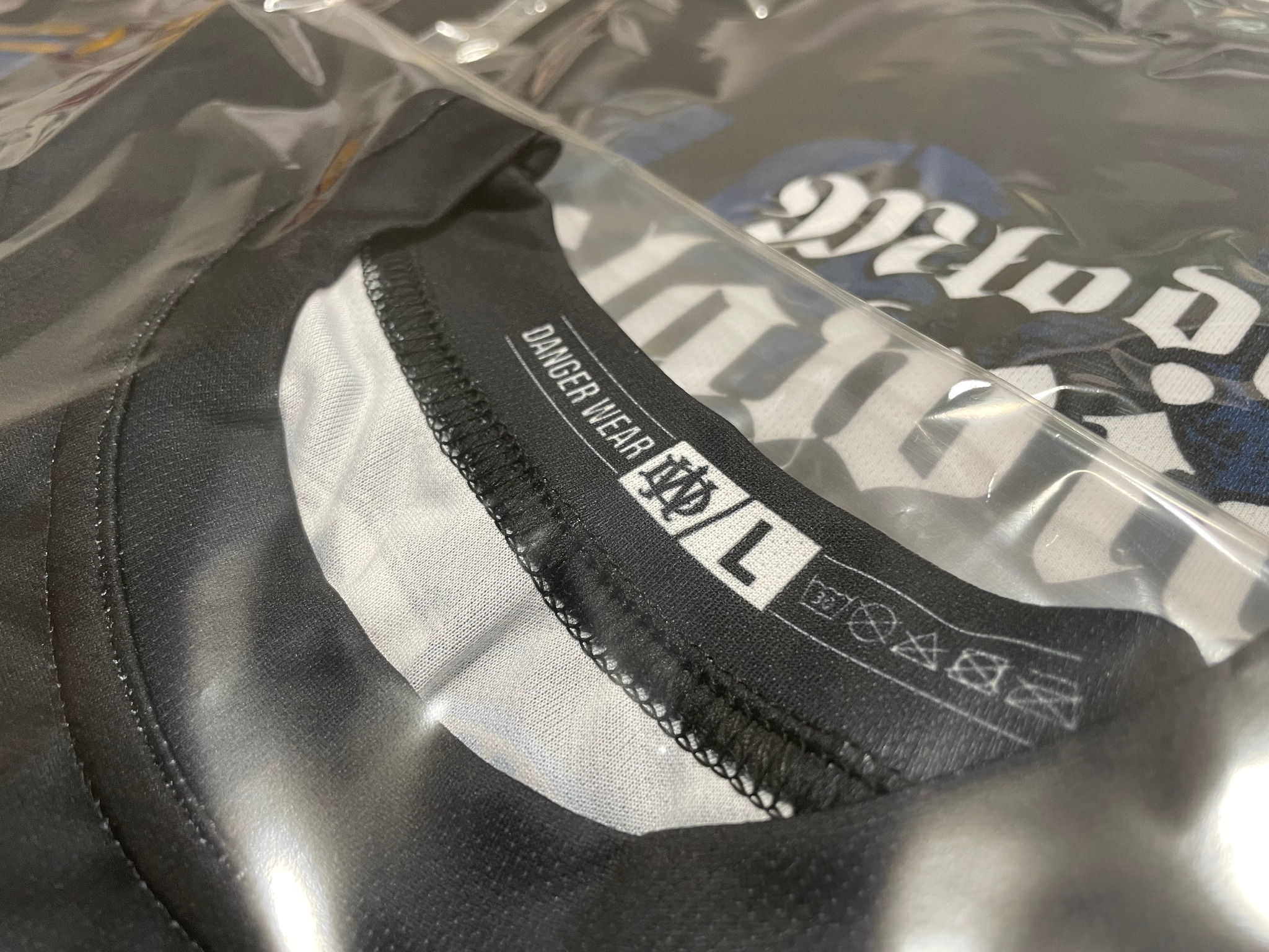 Częściowo rozpakowana czarna koszulka z nadrukiem, widoczna metka 'DANGER WEAR' i rozmiar L, zapakowana w folię.