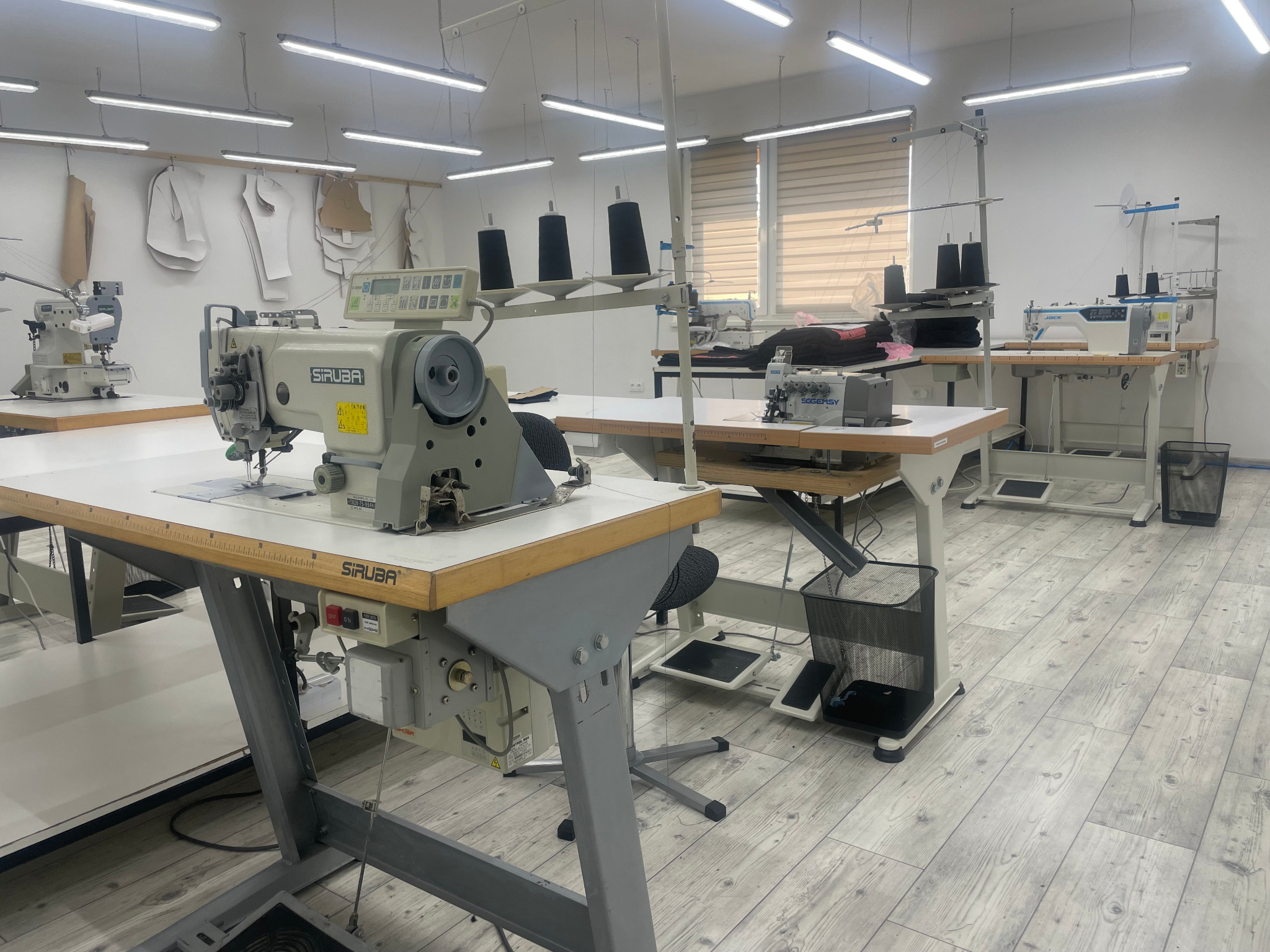 Wnętrze pracowni krawieckiej z kilkoma stanowiskami z maszynami do szycia, widoczne szpulki nici, wykroje odzieży na ścianie i jasna podłoga z paneli.