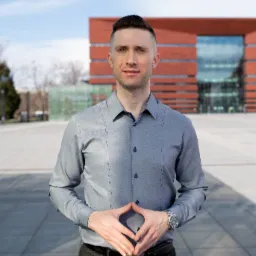 Mężczyzna w szarej koszuli i ciemnych spodniach stoi na placu przed nowoczesnym, ceglanym budynkiem z przeszkloną fasadą, dłonie złożone na brzuchu, widoczny zegarek na nadgarstku.