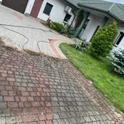 Częściowo wyczyszczona kostka brukowa na podjeździe domu jednorodzinnego, widoczny wąż ciśnieniowy i butelka z detergentem, kontrast między czystą a brudną powierzchnią.