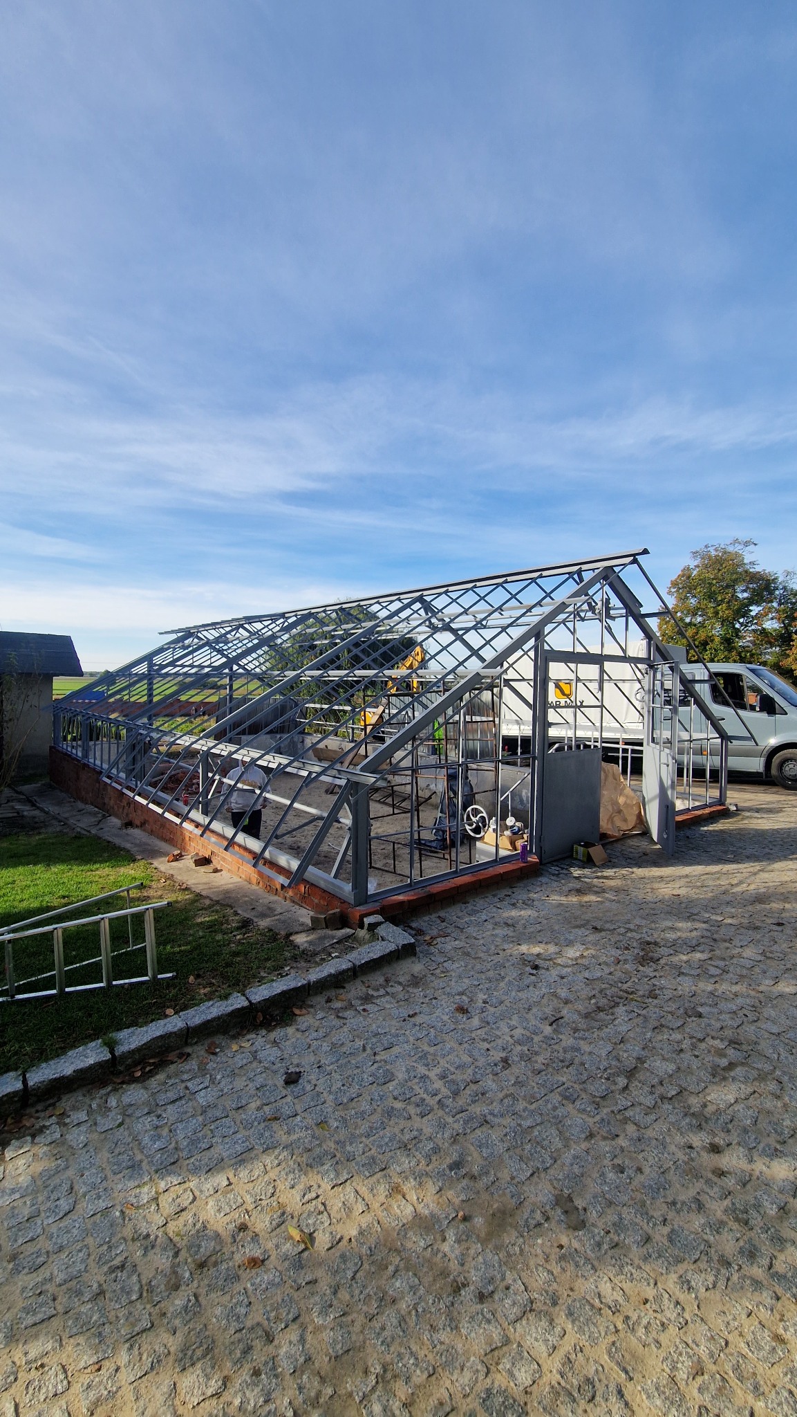 Konstrukcja szkieletowa szklarni z metalu i cegły, częściowo oszklona, na tle błękitnego nieba i zaparkowanego busa.