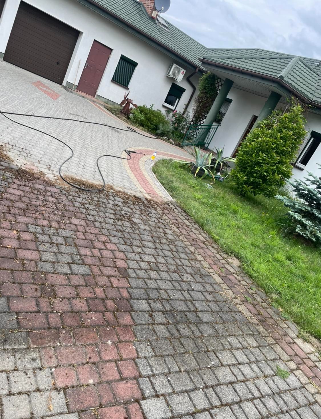 Częściowo wyczyszczona kostka brukowa na podjeździe domu jednorodzinnego, widoczny wąż ciśnieniowy i butelka z detergentem, kontrast między czystą a brudną powierzchnią.