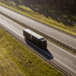 Czarna ciężarówka z naczepą LOGISTIC jadąca autostradą, widok z góry, w tle zielone pola i las.
