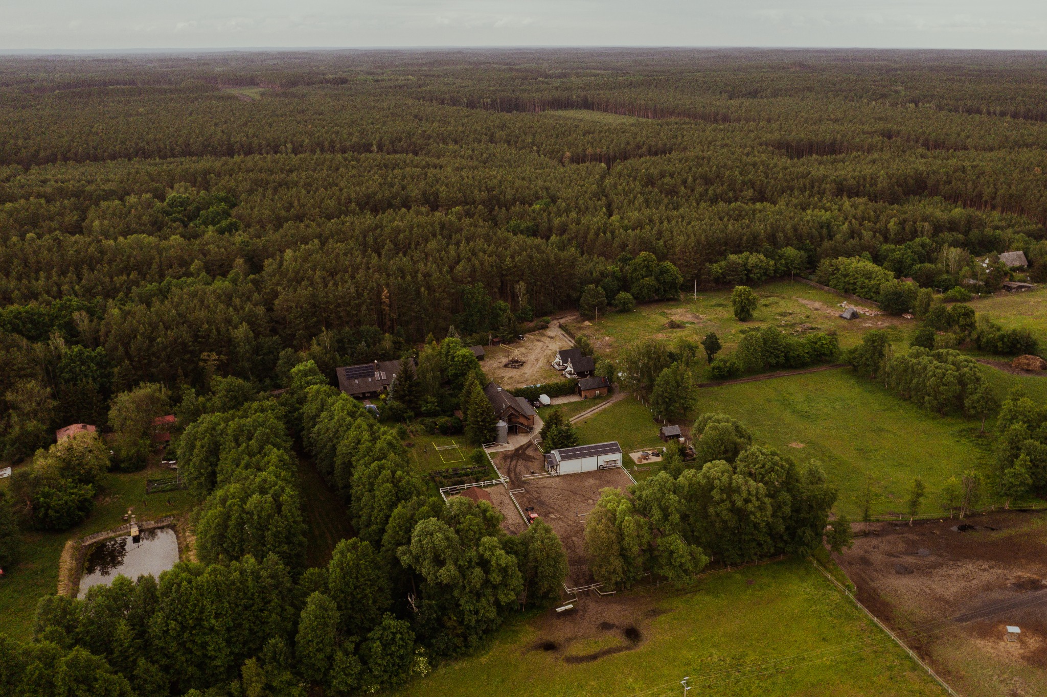 Stajnia Zwierzyniec widziana z lotu ptaka: zabudowania gospodarcze, wybieg dla koni, mały staw otoczony zielenią, w tle rozległy las.