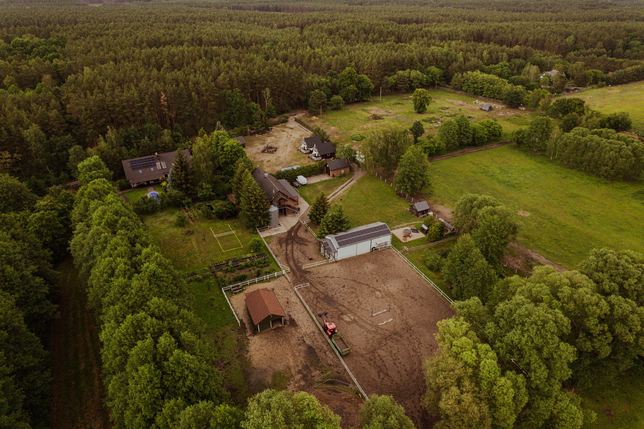 Widok z drona na Stajnię Zwierzyniec: zabudowania gospodarcze, padok z traktorem i przyczepą, otoczone lasem.