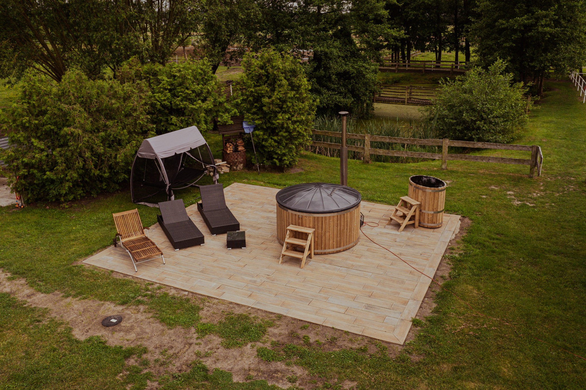 Widok z góry na drewnianą balię z czarną pokrywą i drabinką, oraz drewnianą beczkę z drabinką na tarasie z kostki brukowej w otoczeniu zieleni i drzew w Stajni Zwierzyniec.