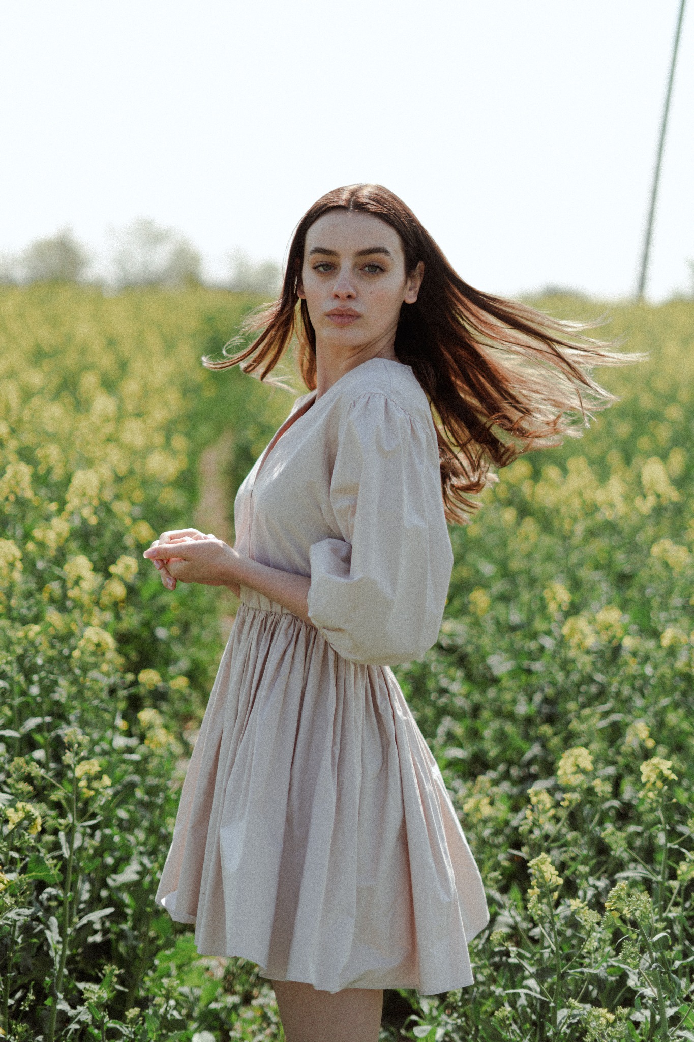 Portret kobiety o długich brązowych włosach w zwiewnej sukience na tle kwitnącego pola rzepaku, włosy w ruchu, spojrzenie w obiektyw.