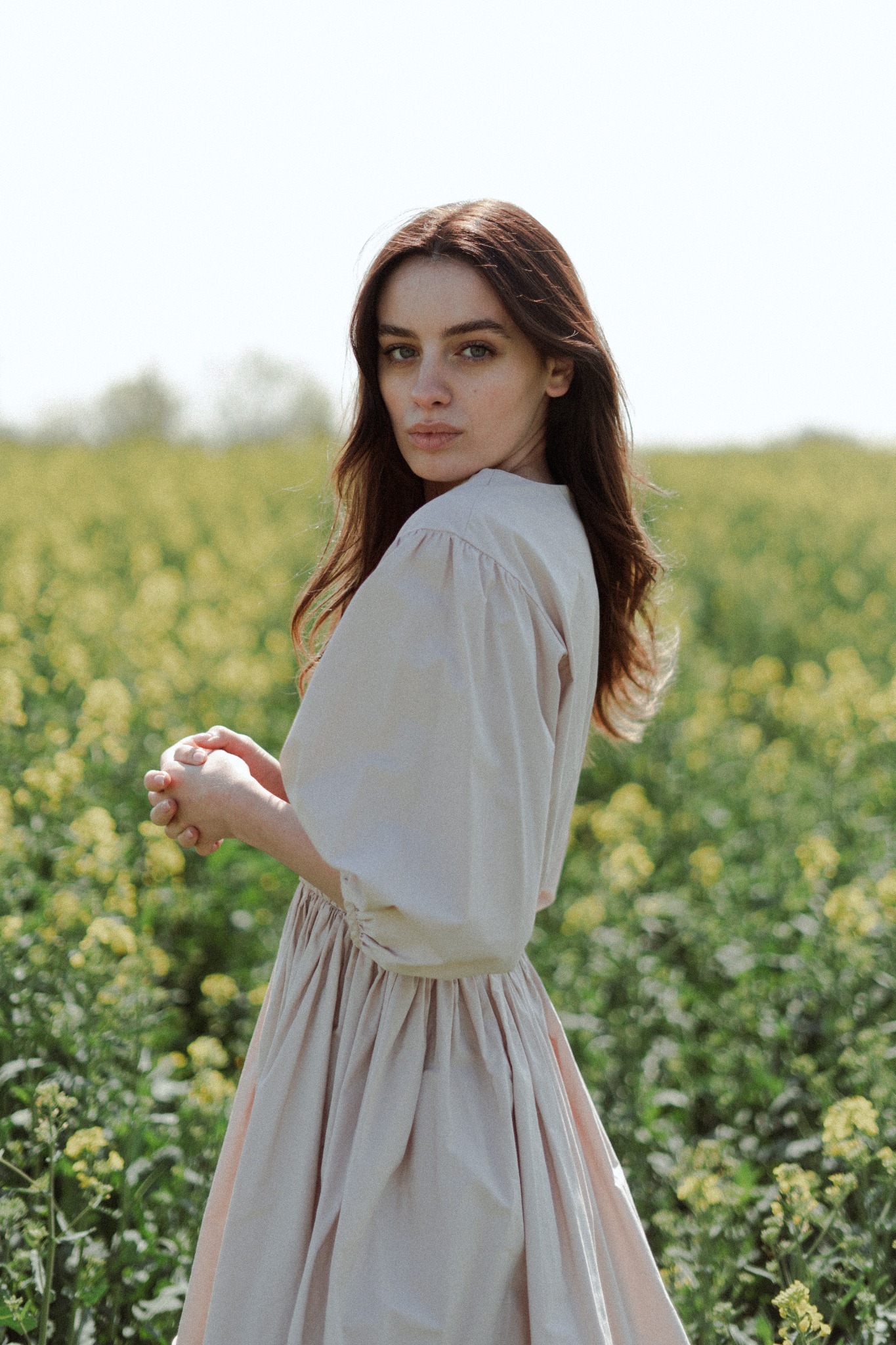 Portret kobiety w zwiewnej, beżowej sukience na tle kwitnącego pola rzepaku, z delikatnym uśmiechem i spojrzeniem przez ramię.