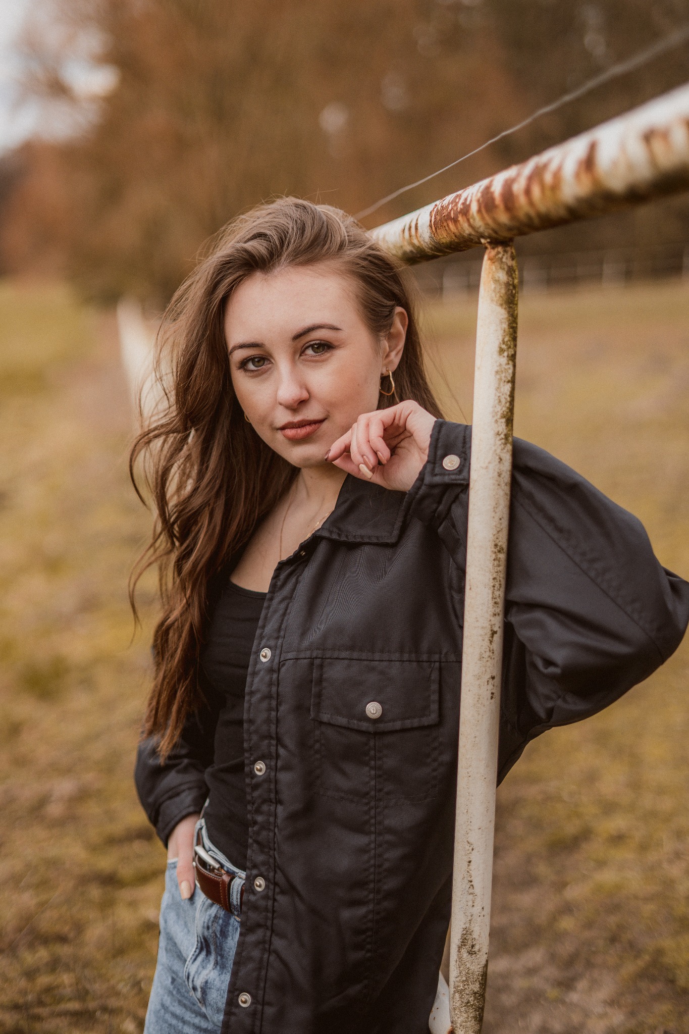 Portret kobiety o długich brązowych włosach, opierającej się o zardzewiałą metalową barierkę w plenerze, ubrana w czarną koszulę i dżinsy.