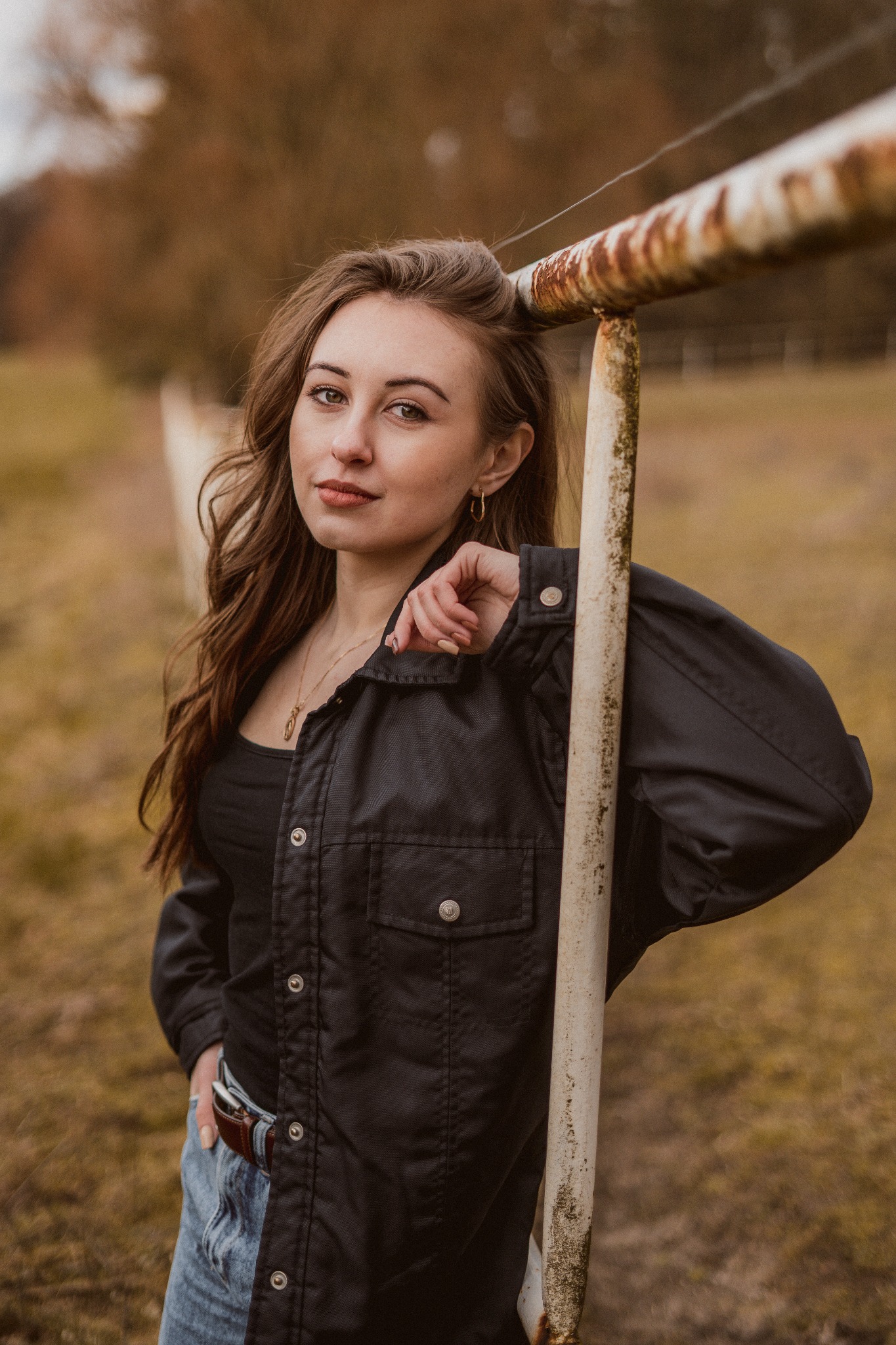 Portret kobiety opierającej się o zardzewiałą metalową barierkę na tle jesiennego krajobrazu, uśmiechająca się, ubrana w czarną kurtkę i dżinsy.
