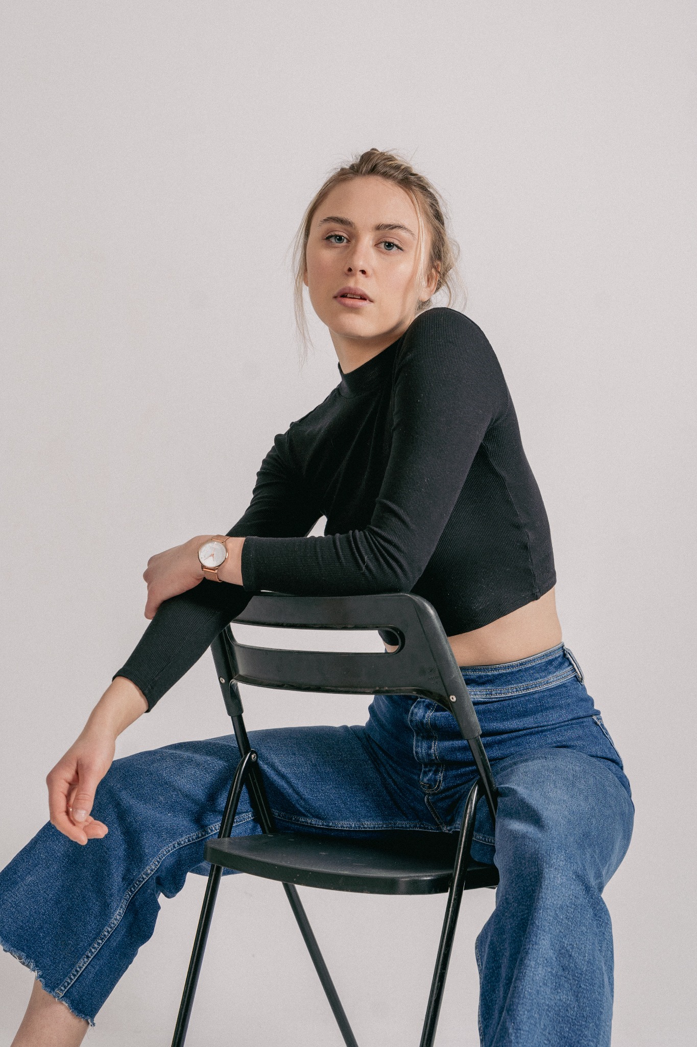Portret młodej kobiety w czarnej bluzce i szerokich jeansach, siedzącej na czarnym krześle na jasnym tle, z zegarkiem na nadgarstku.