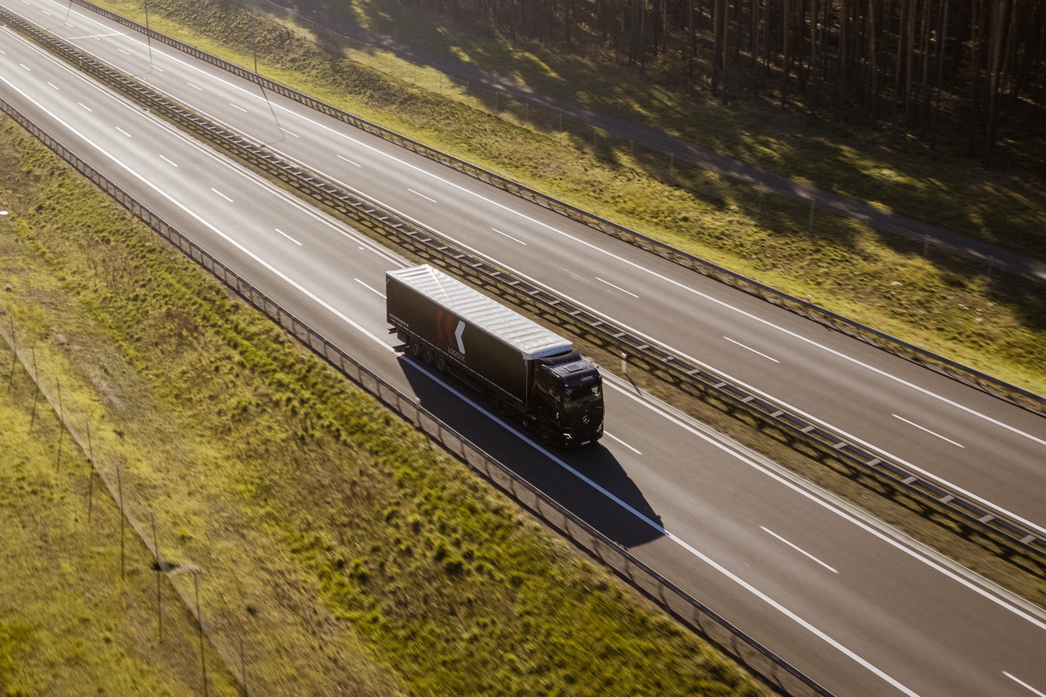 Czarna ciężarówka z naczepą LOGISTIC jadąca autostradą, widok z góry, w tle zielone pola i las.