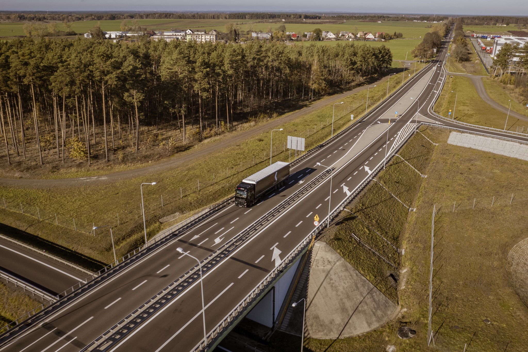 Czarny samochód ciężarowy z naczepą jedzie po estakadzie autostrady, widok z drona, w tle las i zabudowania.