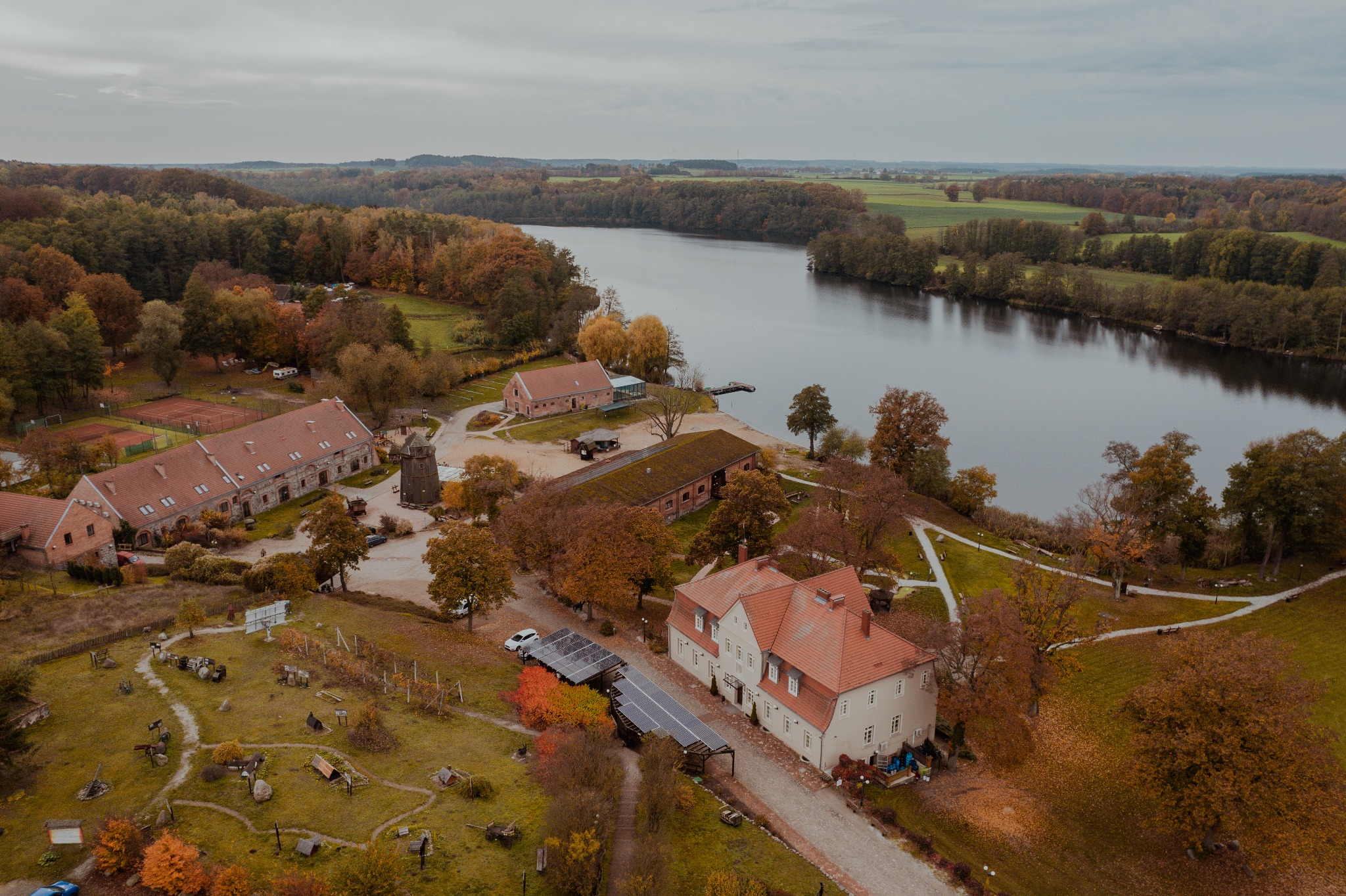 Jesienny krajobraz z lotu ptaka ukazuje kompleks budynków z czerwonymi dachami nad jeziorem, otoczony kolorowymi drzewami i polami uprawnymi pod pochmurnym niebem.