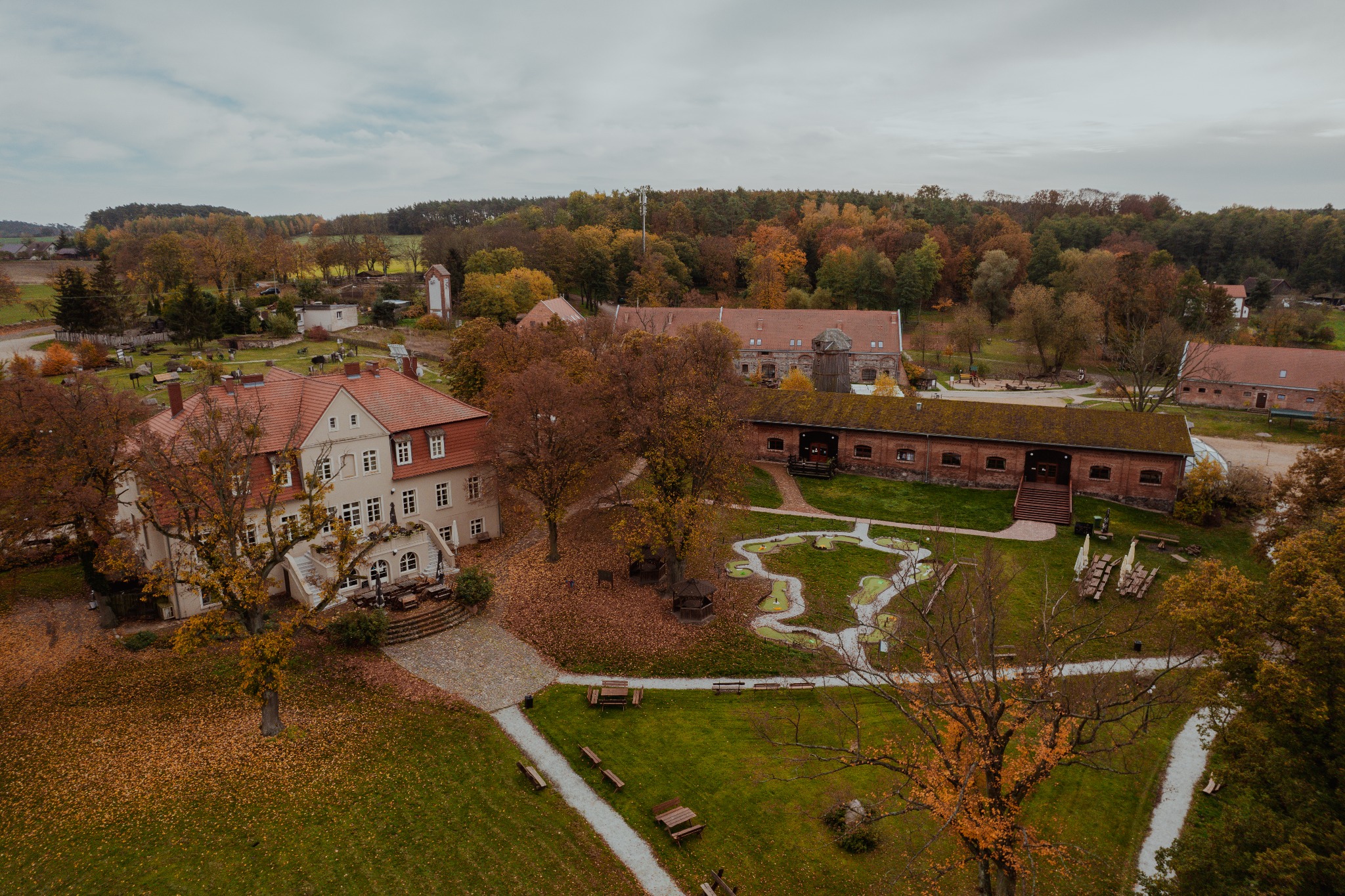 Widok z drona na duży, zabytkowy budynek z czerwonym dachem i parkiem z minigolfem w jesiennej scenerii. Liście pokrywają trawniki i ścieżki.