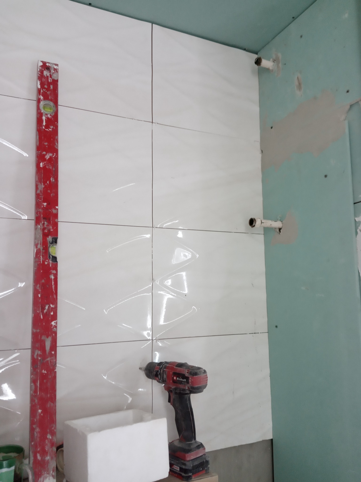 Układanie białych, błyszczących płytek w łazience, widoczny poziomica, wiertarka i płyta gipsowo-kartonowa z wystającymi rurami.