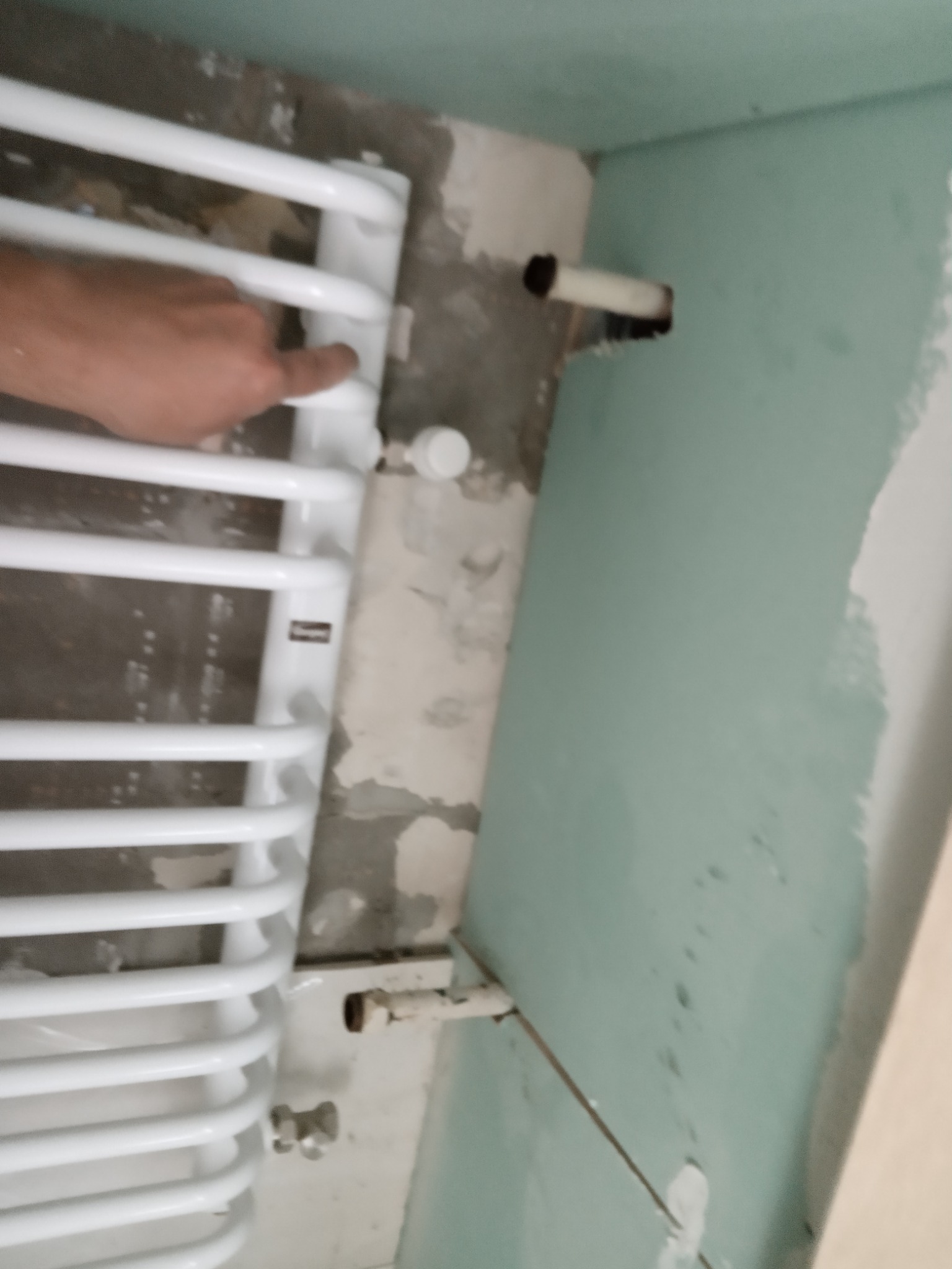 Biały grzejnik łazienkowy, zamontowany na ścianie w trakcie prac wykończeniowych, z widocznymi rurami i fragmentem dłoni wskazującej na element grzejnika.