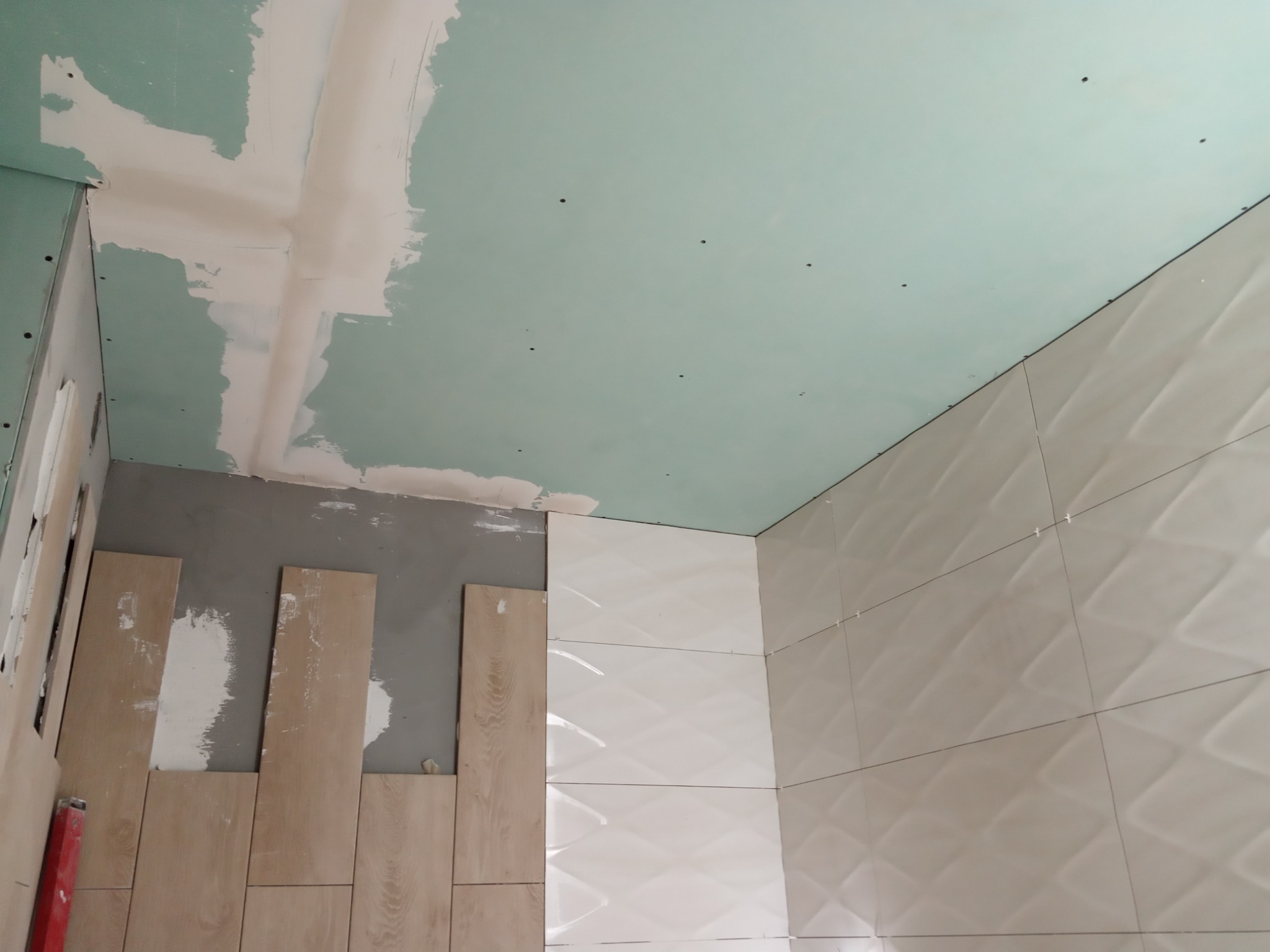 Łazienka w trakcie wykończenia: białe, trójwymiarowe płytki na ścianie, sufit z płyt gipsowo-kartonowych, fragment ściany z przygotowaniem pod płytki drewnopodobne.