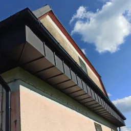 Nowoczesne obróbki blacharskie dachu z czarnej blachy, widoczne rynny i fragment elewacji budynku na tle błękitnego nieba z chmurami.