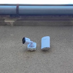 Kamera monitoringu z białą obudową i anteną, zamontowana na szarej, chropowatej elewacji budynku pod rynną.