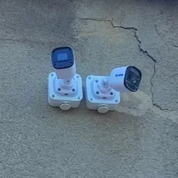 Dwie białe kamery monitoringu zamontowane na szarej, popękanej ścianie zewnętrznej budynku, widoczny fragment rynny.