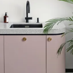 Kuchenny blat z konglomeratu w odcieniach szarości, z czarnym zlewem i baterią, uzupełniony dozownikiem mydła i butelką, na tle pastelowych szafek z okrągłymi, złotymi uchwytami, z dodatkiem...