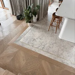 Wnętrze z podłogą łączącą parkiet ułożony w jodełkę i szare płytki imitujące kamień, oddzielone złotą listwą, widoczne schody, robot sprzątający, rośliny doniczkowe i fragment blatu kuchennego.