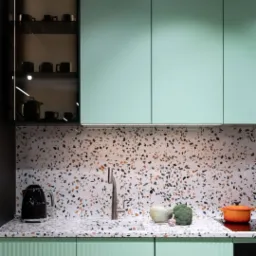 Kuchnia w odcieniach mięty i terrazzo: blat, ściana i fronty szafek z wzorem lastryko, czajnik elektryczny, warzywa i pomarańczowy garnek na płycie indukcyjnej.