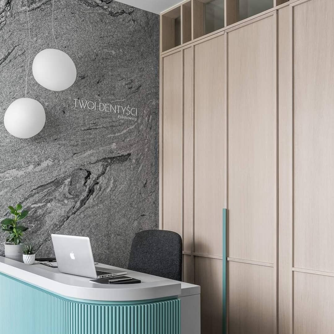 Recepcja w gabinecie dentystycznym z blatem w kolorze turkusowym, komputerem i ścianą z szarego kamienia z napisem 'Twoi Dentyści'.