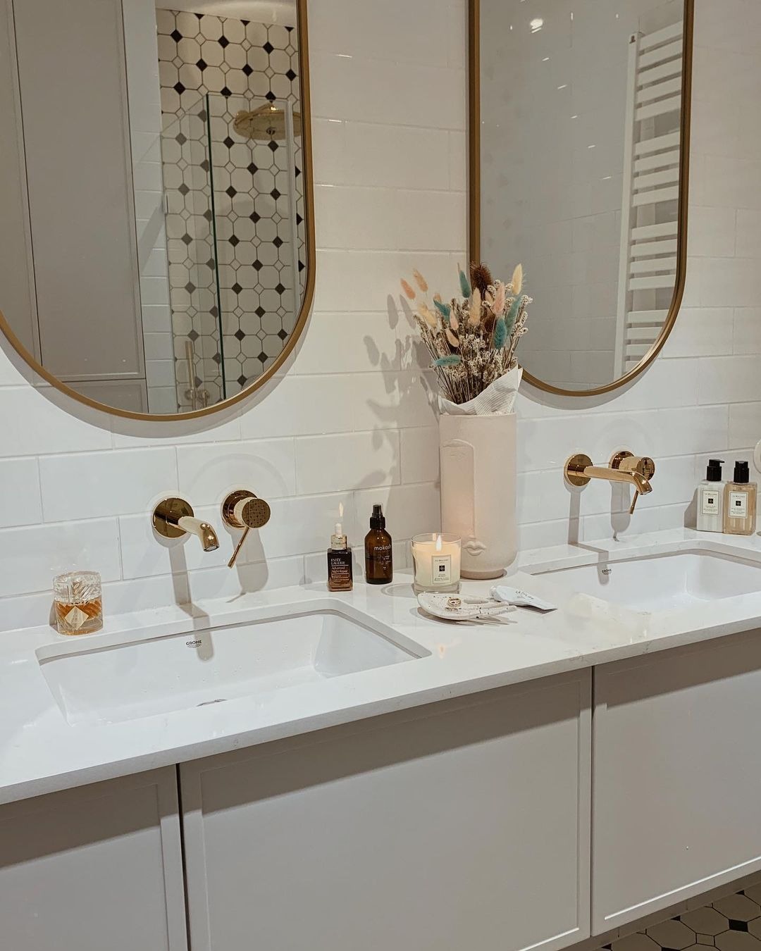 Biała łazienka z dwoma umywalkami nablatowymi, złotą armaturą, lustrami w złotych ramach i dekoracyjnymi detalami, takimi jak suszone kwiaty i świece.