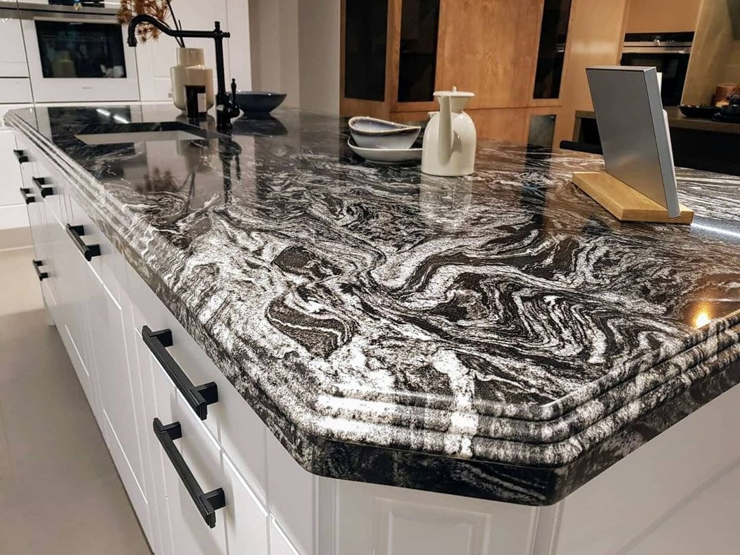 Czarno-biały blat kuchenny z kamienia o unikalnym wzorze żyłkowania, zamontowany na białej wyspie kuchennej z czarnymi uchwytami, widoczne akcesoria kuchenne i oświetlenie.