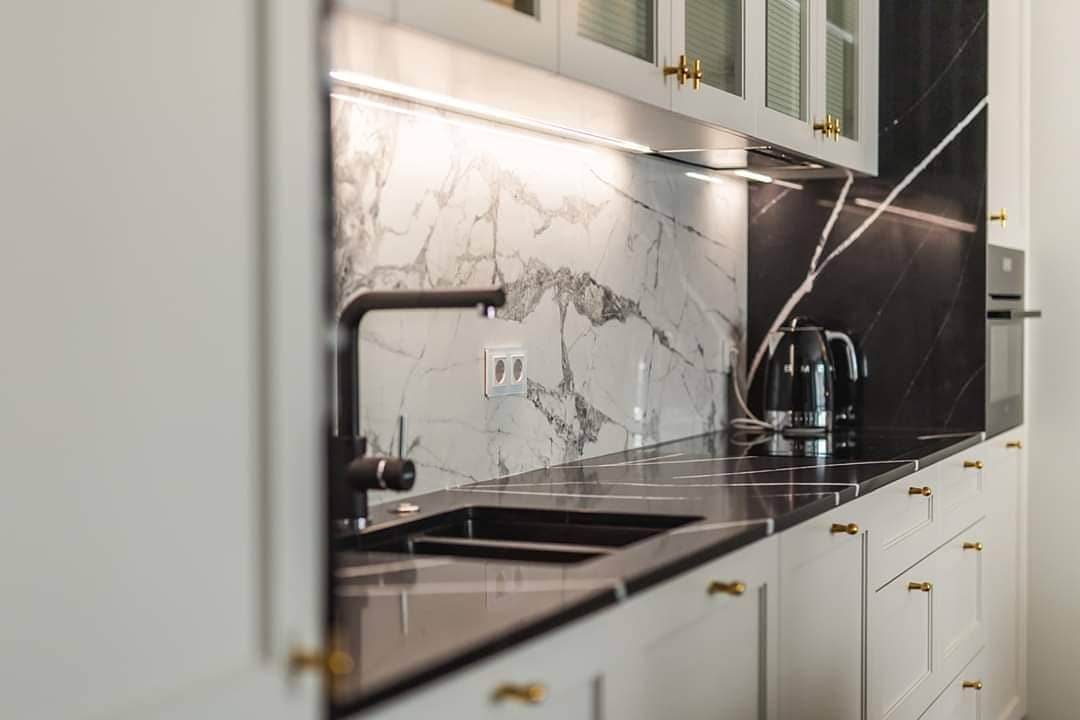 Czarny, błyszczący blat kuchenny z widocznym zlewem i czarną armaturą na tle białej ściany z marmurowym wzorem i szafek z złotymi uchwytami. Na blacie czajnik elektryczny.