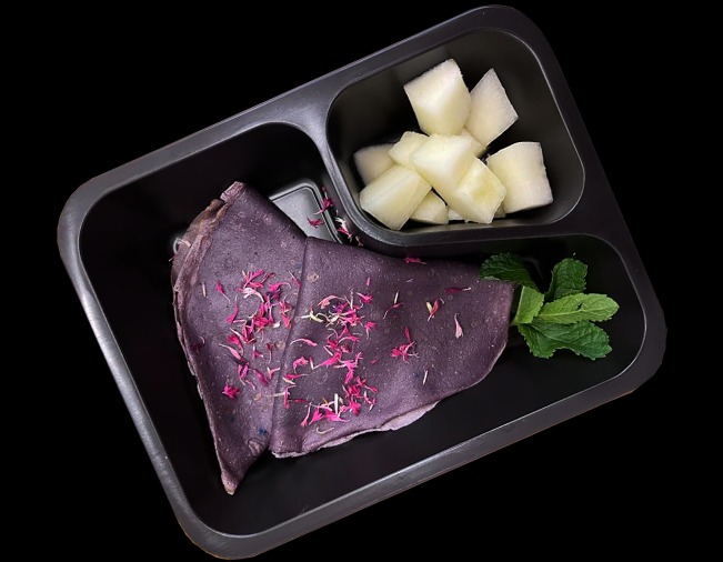 Fioletowe naleśniki posypane drobnymi płatkami kwiatów, kostki melona i listki mięty w czarnym, podzielonym pojemniku na czarnym tle.