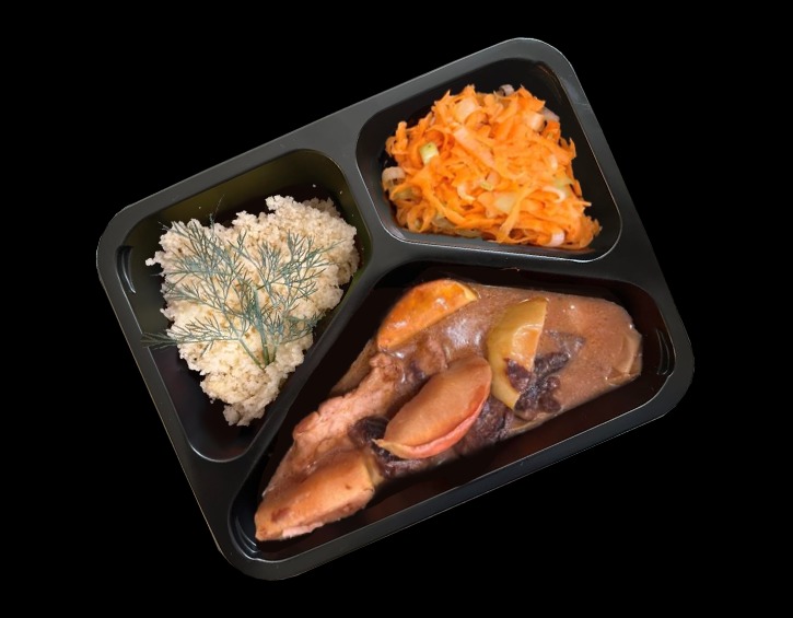 Gotowy posiłek w czarnym opakowaniu z trzema przegródkami: kasza kuskus z koperkiem, surówka z marchewki i mięso duszone z jabłkami i anyżem.