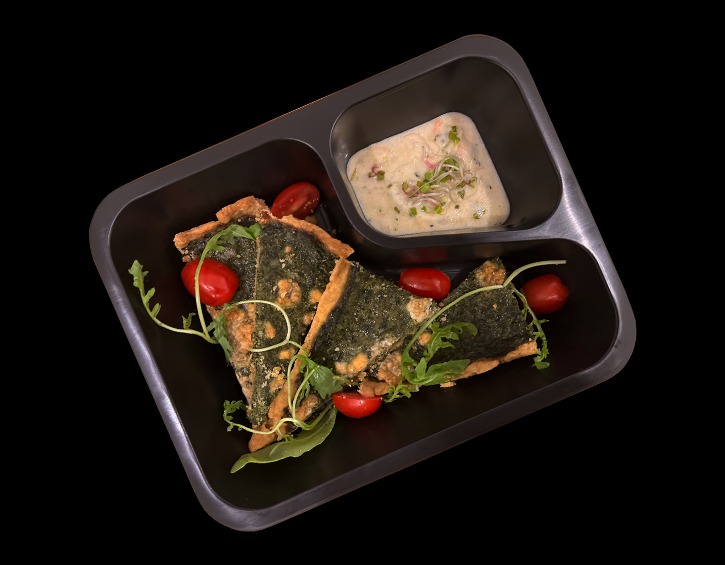 Dwa trójkątne kawałki tarty ze szpinakiem i pomidorkami cherry, podane z sosem w czarnej, dzielonej tacy na czarnym tle.