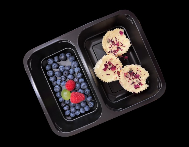 Dwudzielny czarny pojemnik na żywność z borówkami, malinami, zielonym winogronem w jednej przegródce i trzema małymi babeczkami z białym kremem posypanymi płatkami róż w drugiej przegródce,...