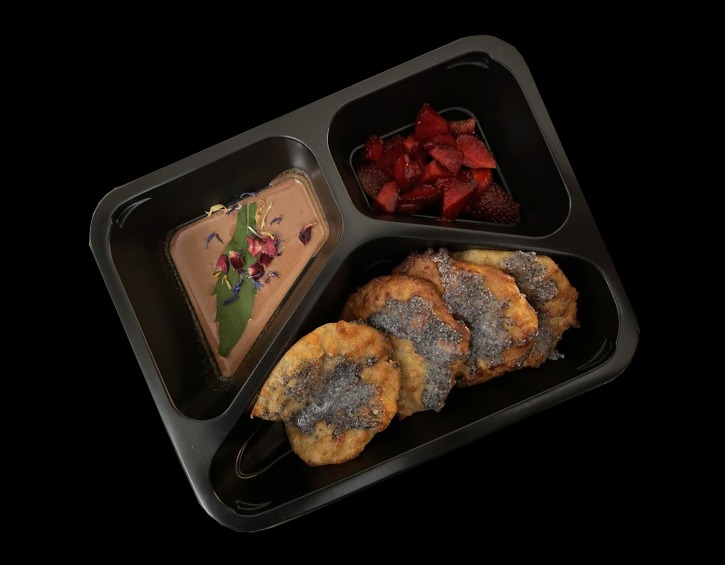 Czarny, trójdzielny pojemnik z jedzeniem: kremowy deser udekorowany kwiatami, pokrojone truskawki i placuszki posypane makiem, na czarnym tle.