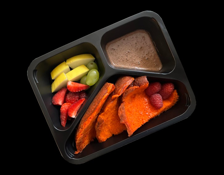 Czarne pudełko z przegródkami zawiera posiłek: w jednej przegródce pokrojone jabłka, winogrona i truskawki, w drugiej brązowy krem, a w trzeciej pomarańczowe tosty francuskie z malinami na wierzchu.