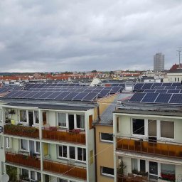 Bpower - Widok z góry na dachy budynków mieszkalnych z zainstalowanymi panelami słonecznymi; dwóch mężczyzn stoi na dachu podczas instalacji lub konserwacji, na tle miejski krajobraz z wieżowcem...