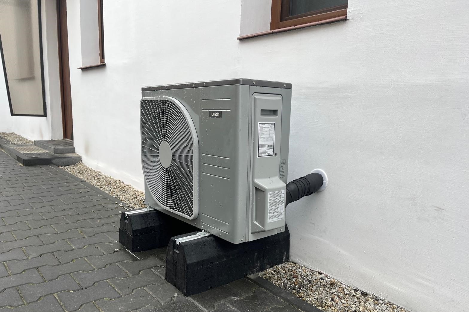 Szara jednostka zewnętrzna pompy ciepła L6split zamontowana na czarnych, betonowych blokach przy białej ścianie budynku z widocznym przewodem odprowadzającym skropliny.