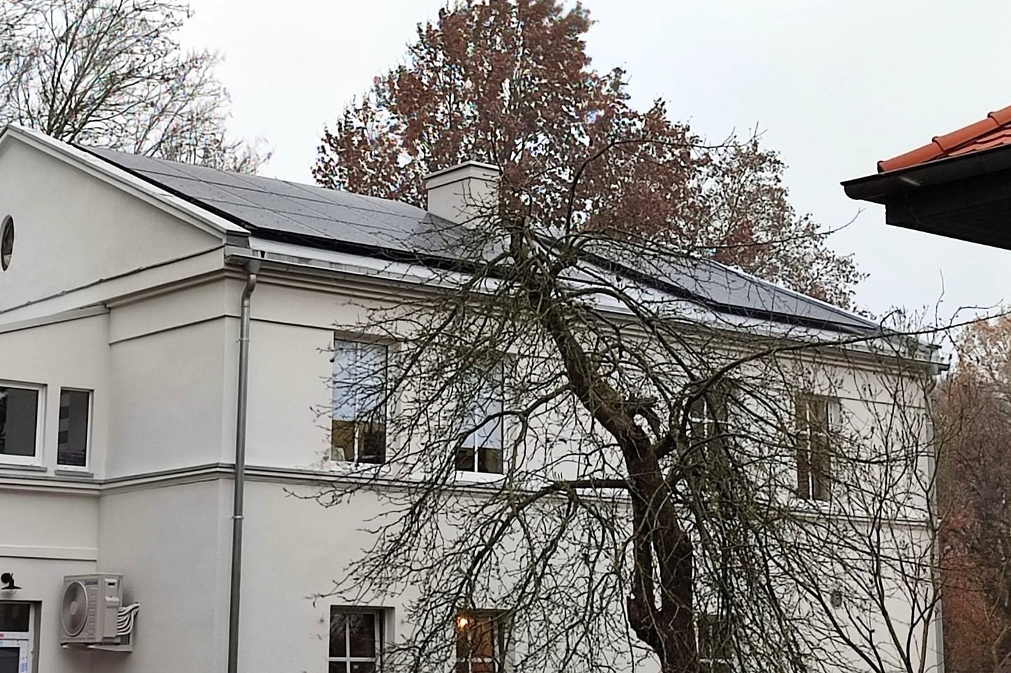 Biały budynek z panelami słonecznymi na dachu, widziany przez gałęzie drzewa bez liści i z kominem w tle.