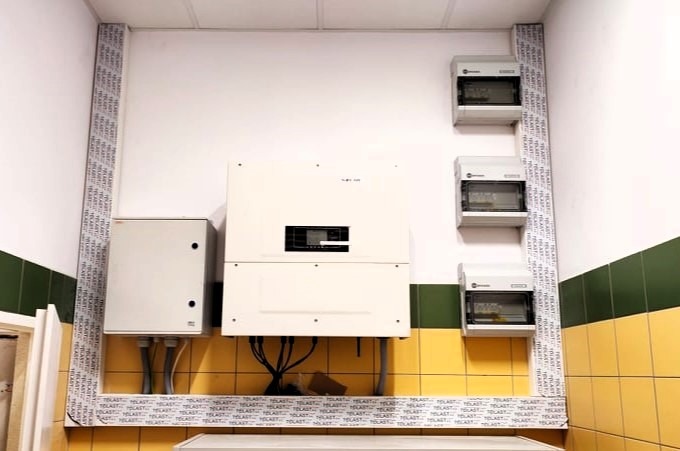 Biała ściana z zamontowanymi: skrzynką elektryczną, inwerterem i trzema rozdzielnicami, zabezpieczona profilem z napisem 'TELAST'.