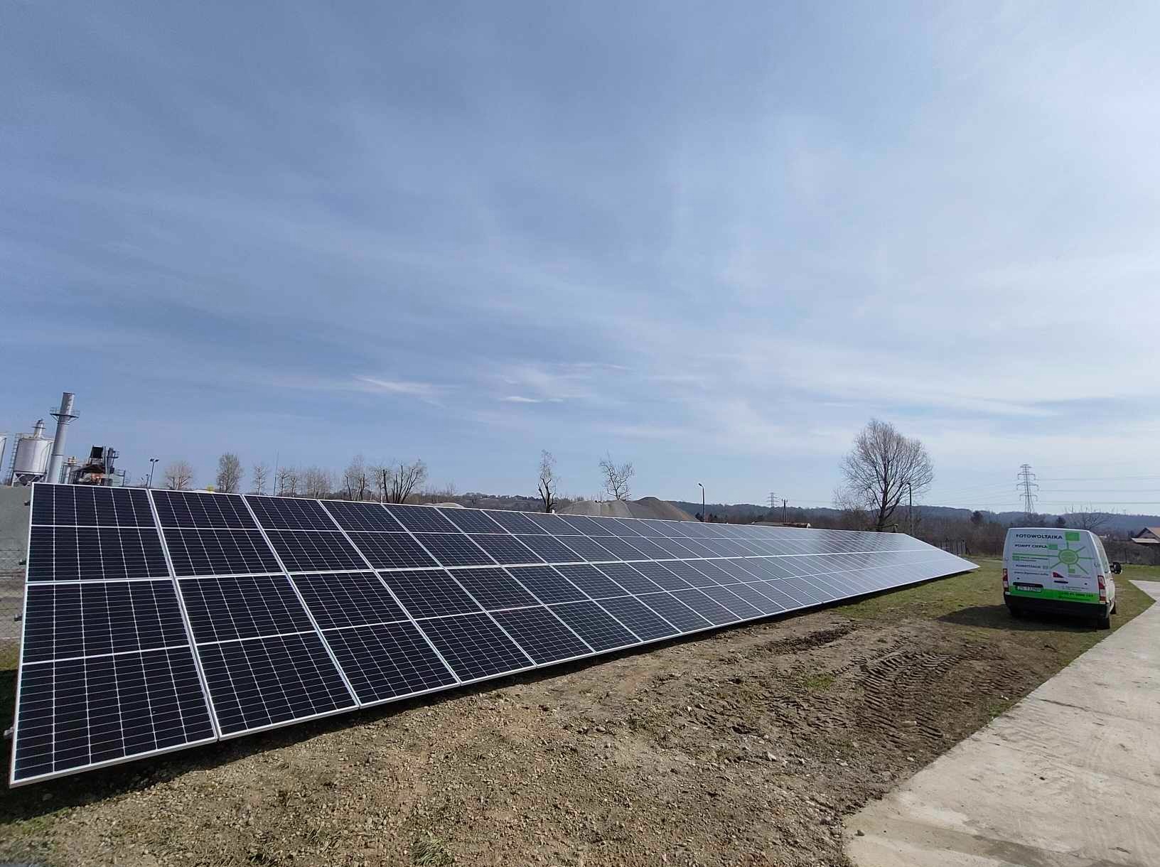 Rozległa instalacja paneli słonecznych na gruncie, z widocznym samochodem dostawczym firmy instalacyjnej w tle, na tle błękitnego nieba.