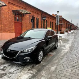 Czarna taksówka Mazda zaparkowana na brukowanej ulicy w tle ceglane budynki z szyldami Żabka i Sugar.