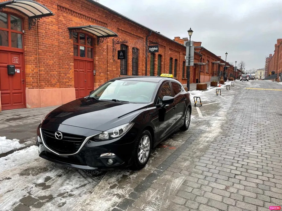 Czarna taksówka Mazda zaparkowana na brukowanej ulicy w tle ceglane budynki z szyldami Żabka i Sugar.