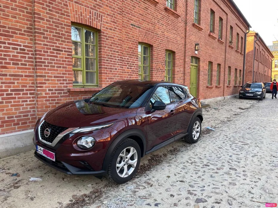 Bordowy Nissan Juke zaparkowany na brukowanej ulicy w Łodzi na tle ceglanego budynku z zielonymi oknami.