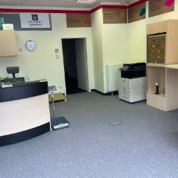 Wnętrze punktu usługowego z ladą recepcyjną, wagą, drukarką i ścianą z przegródkami pocztowymi. Widoczne plakaty informujące o usługach pocztowych, e-usługach, spedycji i druku cyfrowym.