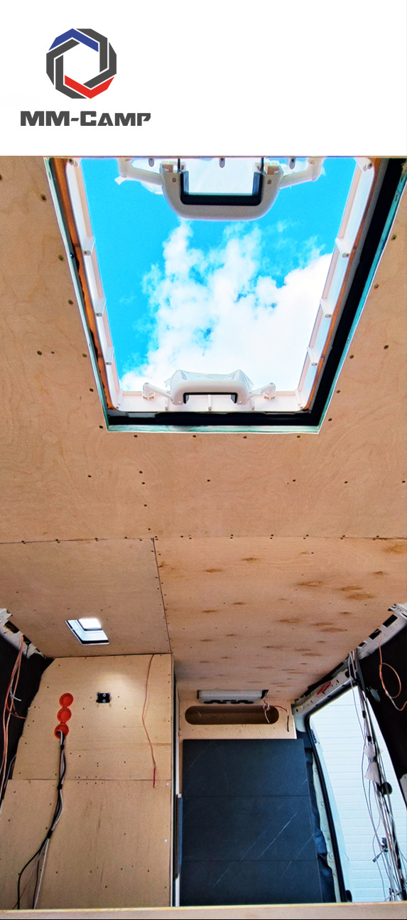 Wnętrze furgonetki w trakcie adaptacji: drewniana boazeria, okno dachowe z widokiem na błękitne niebo, instalacja elektryczna w trakcie montażu, widoczny fragment zabudowy.