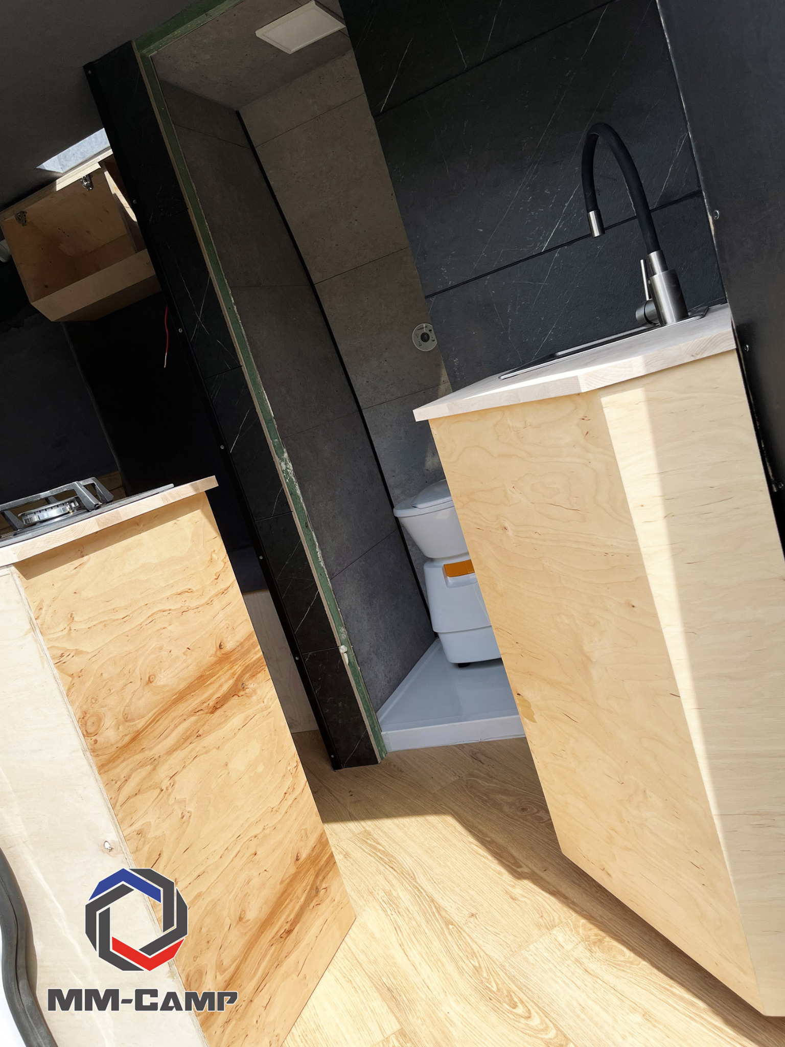 Wnętrze kampera w trakcie zabudowy, widoczne drewniane meble kuchenne z blatem i zlewem z czarną armaturą, obok toaleta turystyczna w kabinie prysznicowej, logo firmy MM-CAMP w rogu zdjęcia.